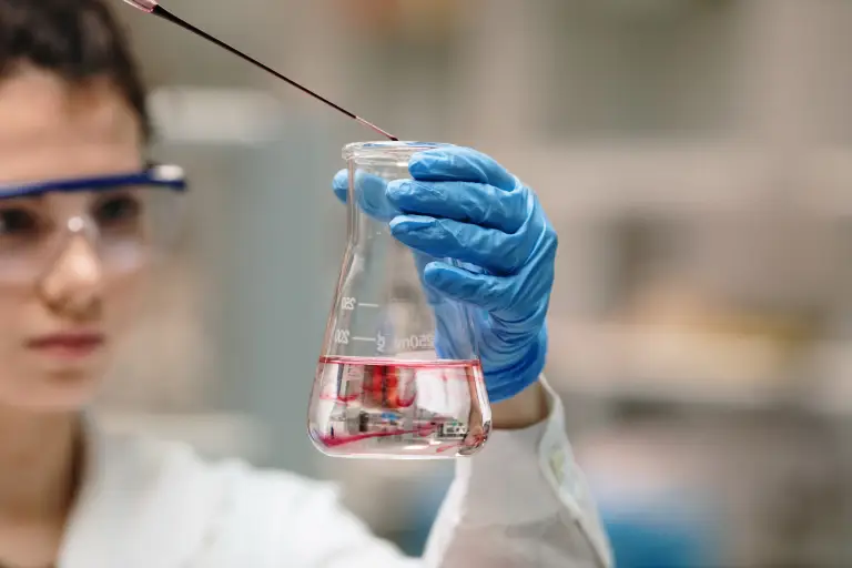 Person in safety goggles and blue lab gloves adding drops of red liquid to a conical flask