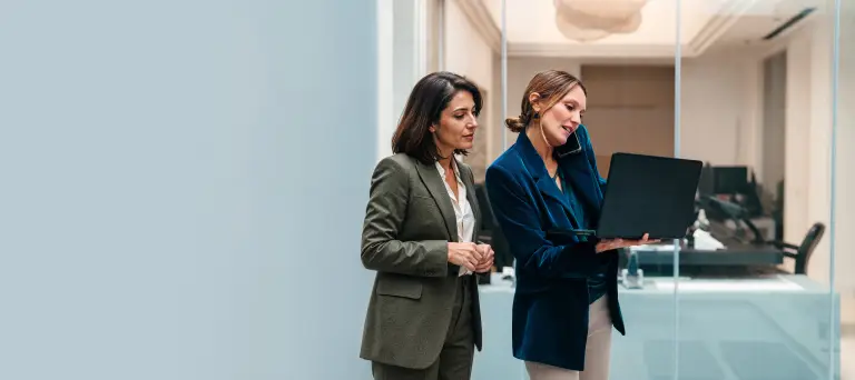 Zwei Frauen in Businesskleidung besprechen sich vor einem Laptop in einem Büro mit Glaswänden